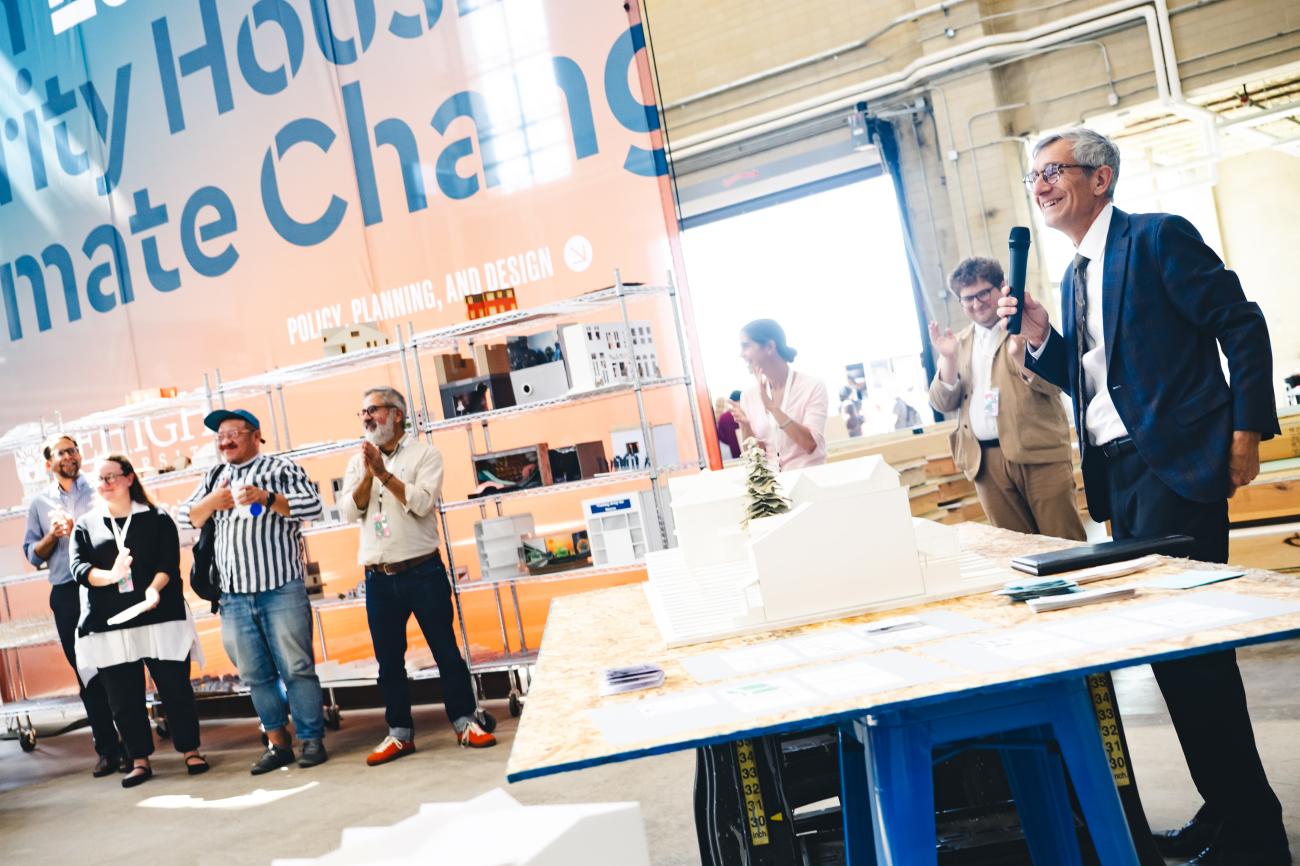 President Helble standing in front of 3D models of houses talking to an audience with banner showing policy, planning, and design.